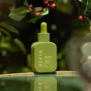 Green skincare bottle labeled '9SKIN' on a reflective surface with blurred greenery in the background