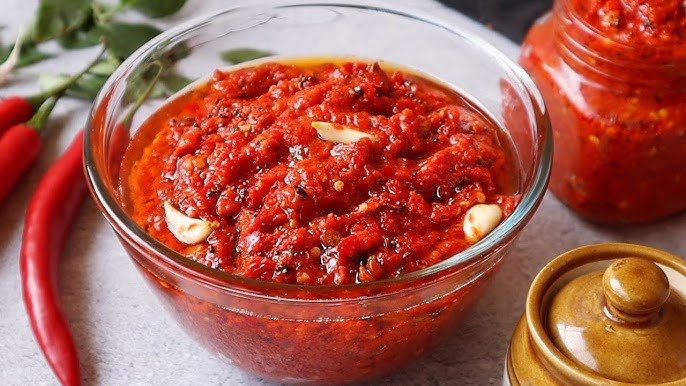 Red chili paste in a glass bowl with red chilies and a jar in the background.