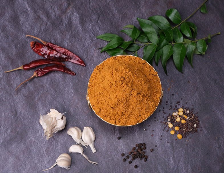 Spice blend in a bowl with red chilies, garlic, and green leaves on a dark surface
