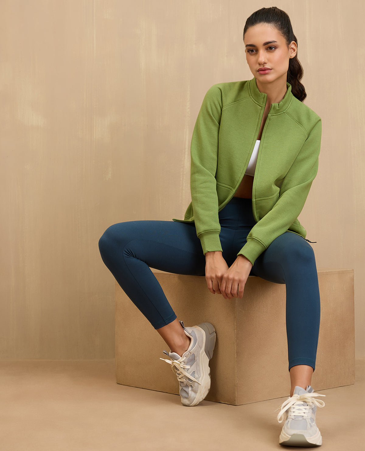 Woman wearing a green jacket and blue leggings sitting on a cardboard box against a beige background