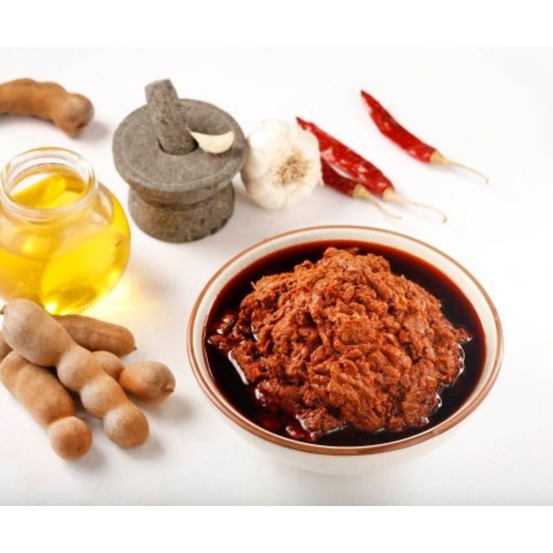Bowl of red paste with tamarind pods, oil, and spices on a white background