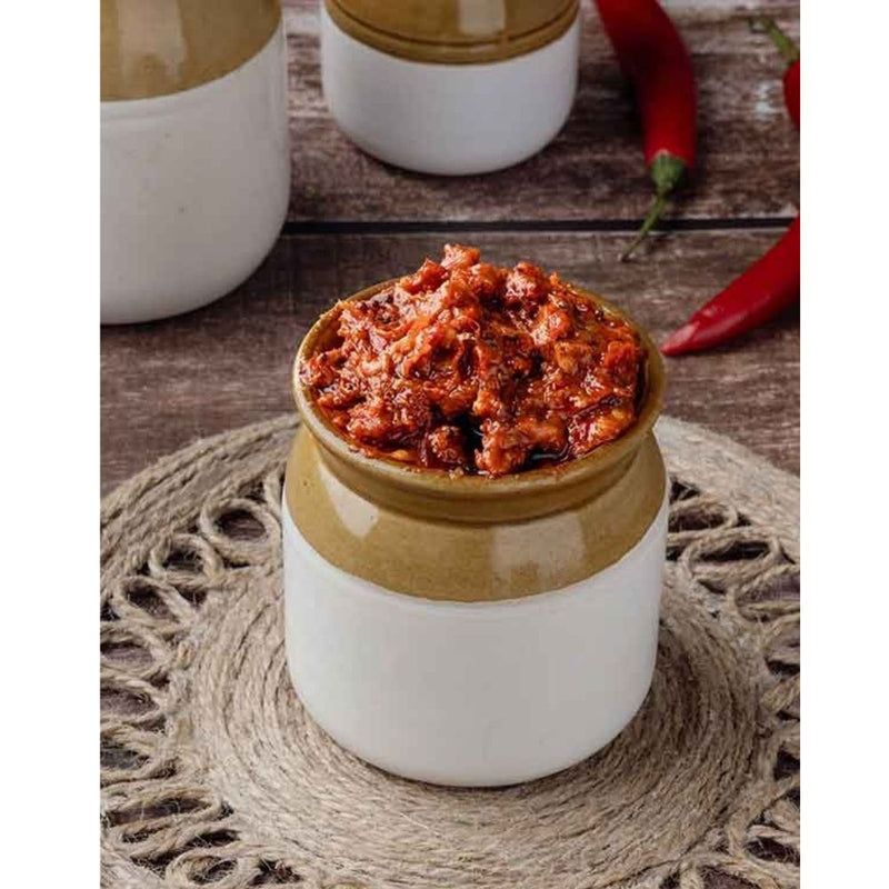 Jar of spicy-looking food on a woven mat with red peppers in the background