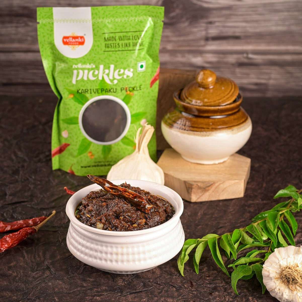 Green packet of pickles with a bowl of pickle, garlic, and red chili peppers on a dark surface.